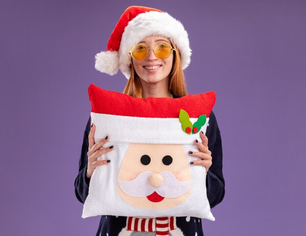 Sonriente joven hermosa niña vistiendo suéter de Navidad y sombrero con gafas sosteniendo almohada de Navidad aislado sobre fondo púrpura