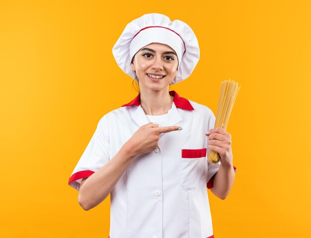 Sonriente joven hermosa en uniforme de chef sosteniendo y puntos en espaguetis