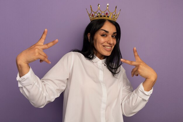 Sonriente joven hermosa vestida con camiseta blanca y corona mostrando gesto de paz aislado en púrpura