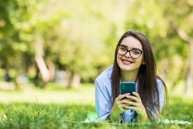 Sonriente joven hipster chica con teléfono inteligente tirado en el césped en el parque