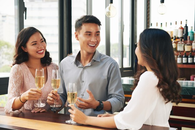 Sonriente joven hombre asiático y dos mujeres animando con champán en el bar