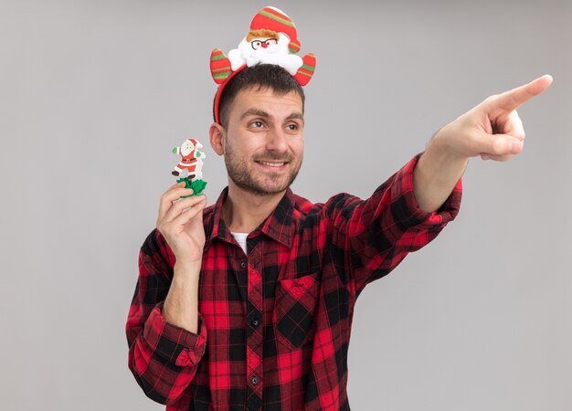 Sonriente joven hombre caucásico con diadema de santa claus sosteniendo muñeco de nieve juguete de Navidad mirando y apuntando al lado aislado sobre fondo blanco.