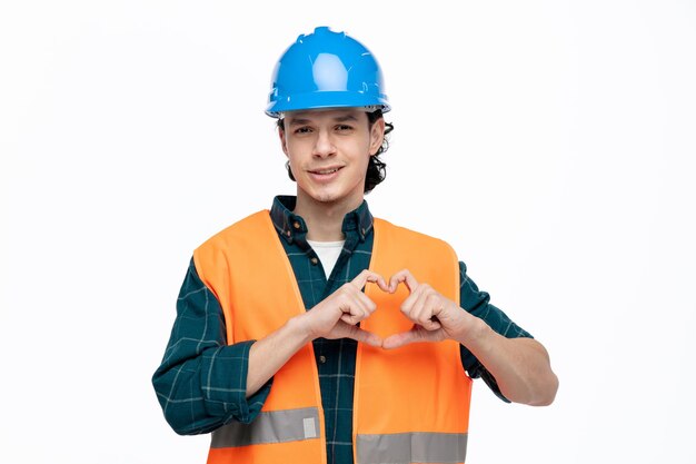 Sonriente joven ingeniero masculino con casco de seguridad y chaleco de seguridad mirando a la cámara mostrando el signo de amor aislado en el fondo blanco