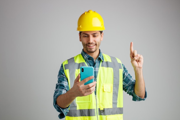 Sonriente joven ingeniero masculino con casco de seguridad y uniforme sosteniendo y mirando el teléfono móvil apuntando hacia arriba aislado en fondo blanco