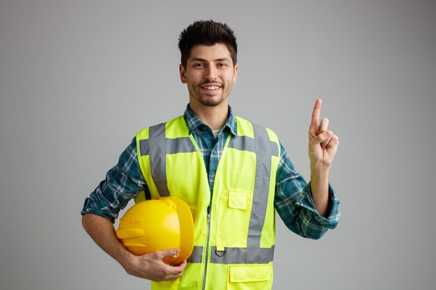 Sonriente joven ingeniero masculino vistiendo uniforme sosteniendo casco de seguridad mirando a la cámara apuntando hacia arriba aislado sobre fondo blanco.