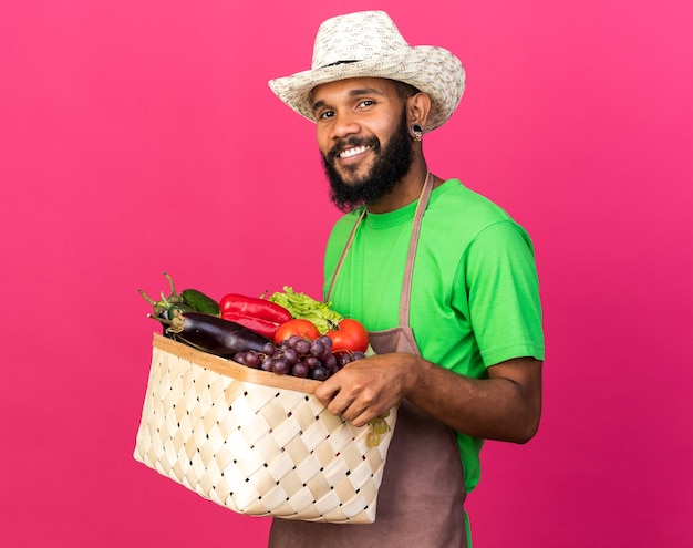 Sonriente joven jardinero afroamericano vistiendo sombrero de jardinería con canasta de verduras aislado en la pared rosa