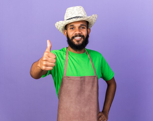 Sonriente joven jardinero afroamericano vistiendo sombrero de jardinería mostrando el pulgar hacia arriba