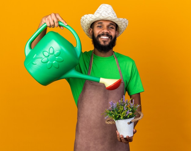 Foto gratuita sonriente joven jardinero afroamericano vistiendo sombrero de jardinería regar las flores en maceta con regadera