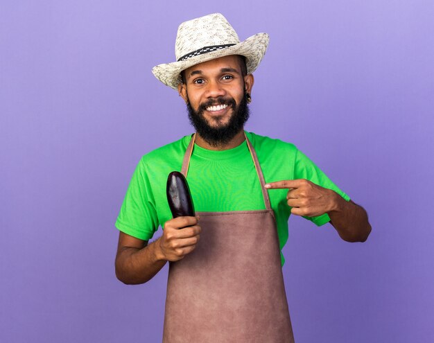 Sonriente joven jardinero afroamericano vistiendo sombrero de jardinería sosteniendo y apunta a la berenjena