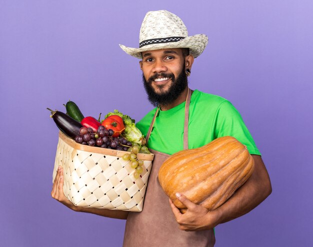 Sonriente joven jardinero afroamericano vistiendo sombrero de jardinería sosteniendo calabaza con canasta de verduras
