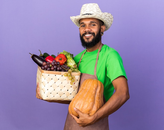 Sonriente joven jardinero afroamericano vistiendo sombrero de jardinería sosteniendo canasta de verduras con calabaza