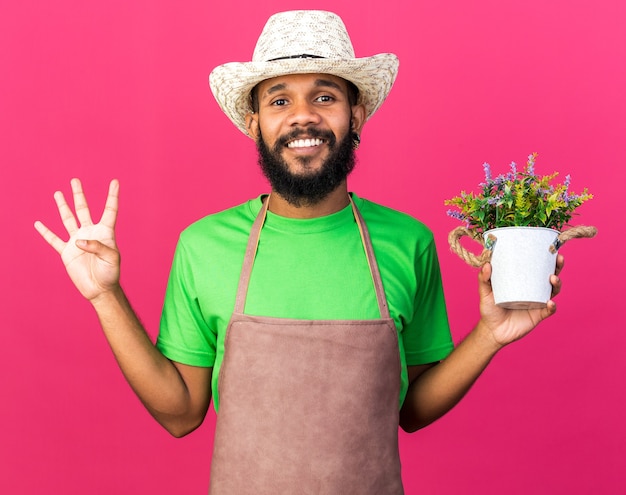 Sonriente joven jardinero afroamericano vistiendo sombrero de jardinería sosteniendo flor en maceta mostrando cuatro aislados en la pared rosa