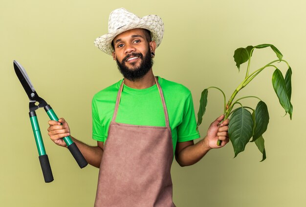 Sonriente joven jardinero afroamericano vistiendo sombrero de jardinería sosteniendo plante con tijeras aisladas sobre pared verde oliva