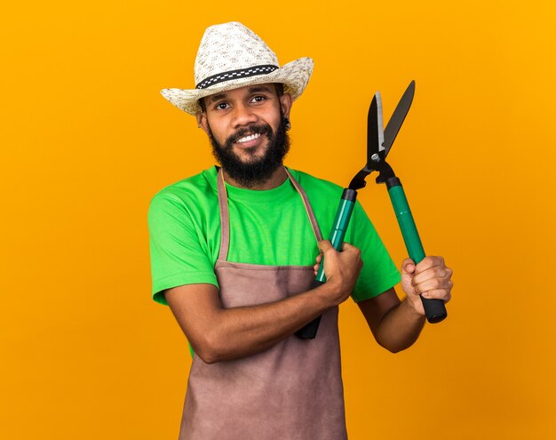 Sonriente joven jardinero afroamericano vistiendo sombrero de jardinería sosteniendo tijeras aisladas sobre pared naranja