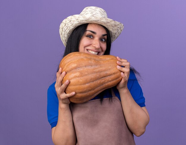 Sonriente joven jardinero caucásico vestida con uniforme y sombrero sosteniendo calabaza butternut aislado en la pared púrpura