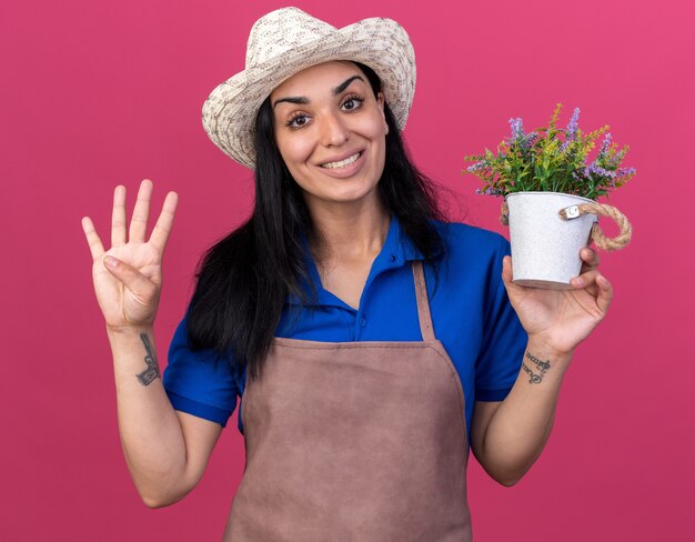 Sonriente joven jardinero caucásico vestida con uniforme y sombrero sosteniendo maceta mostrando cuatro con la mano aislada en la pared rosa