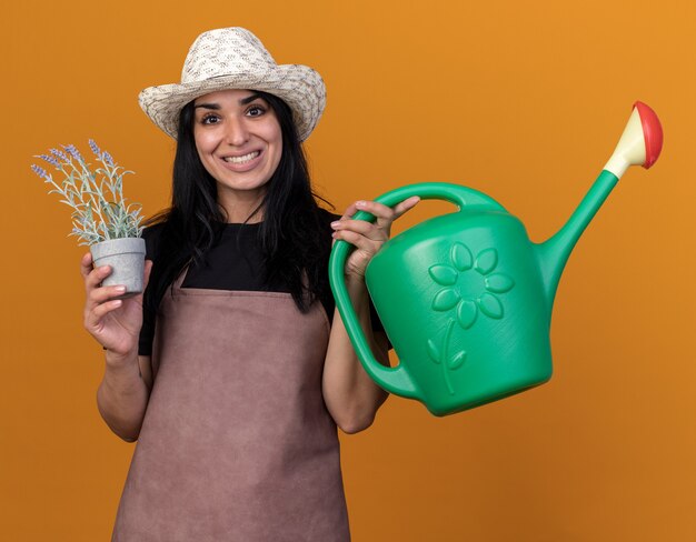 Sonriente joven jardinero caucásico vestida con uniforme y sombrero sosteniendo maceta y regadera