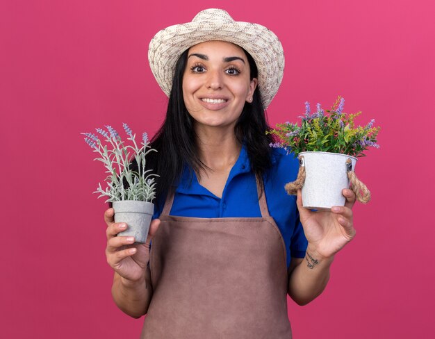 Sonriente joven jardinero caucásico vestida con uniforme y sombrero sosteniendo macetas