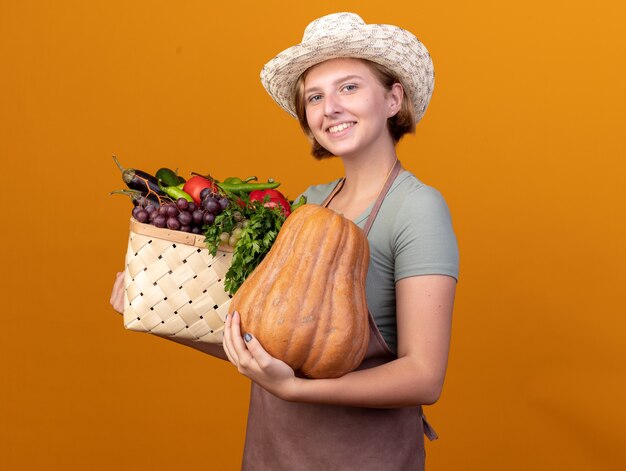 Sonriente joven jardinero eslavo vistiendo sombrero de jardinería sosteniendo canasta de verduras y calabaza en naranja