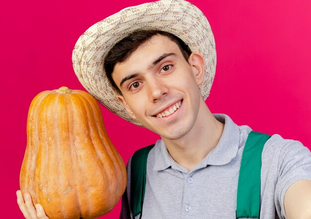 Sonriente joven jardinero con sombrero de jardinería sostiene calabaza y pretende sostener la cámara aislada sobre fondo rosa con espacio de copia