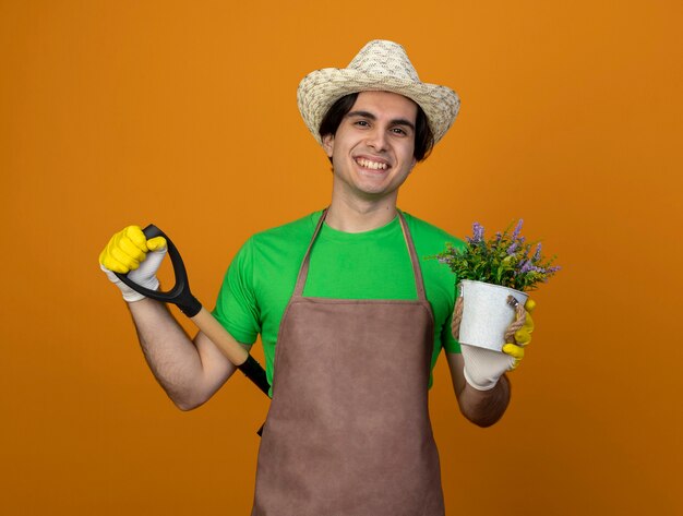 Sonriente joven jardinero en uniforme con sombrero de jardinería con guantes sosteniendo flor en maceta poniendo pala en la cintura aislada en naranja