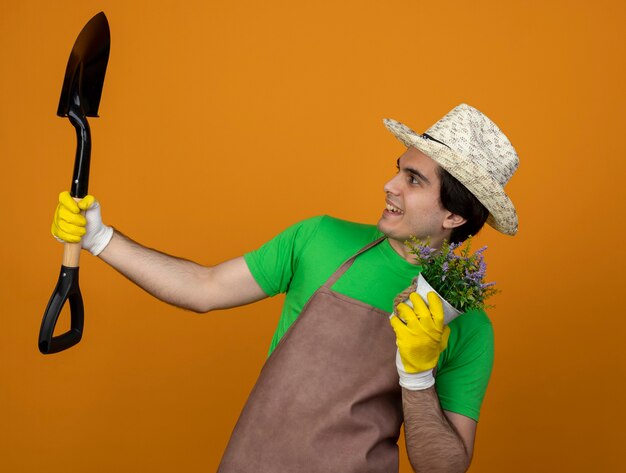 Sonriente joven jardinero en uniforme vistiendo gorro de jardinería con guantes sosteniendo flor en maceta y levantando y mirando a pala