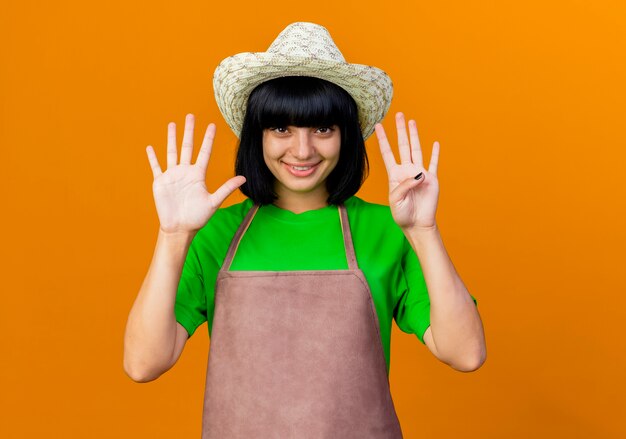 Sonriente joven jardinero en uniforme vistiendo sombrero de jardinería gestos nueve con los dedos