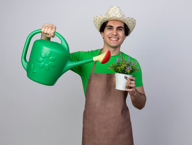 Sonriente joven jardinero en uniforme vistiendo sombrero de jardinería regar las flores en maceta con regadera