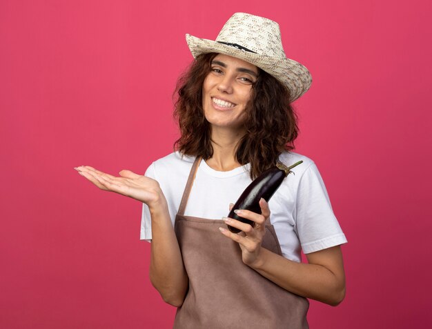 Sonriente joven jardinero en uniforme vistiendo sombrero de jardinería sosteniendo berenjena extendiendo la mano