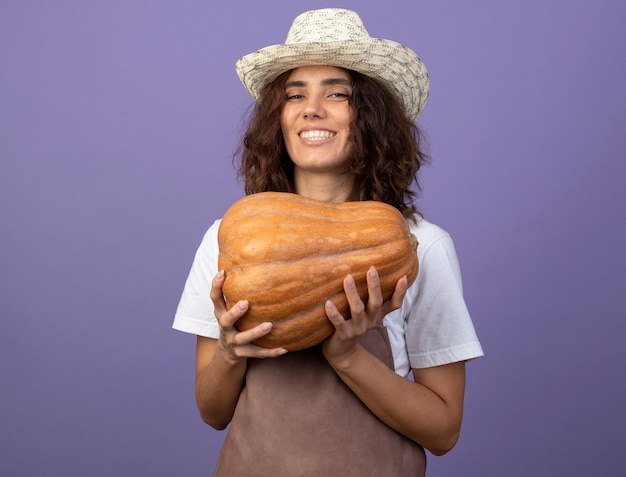 Sonriente joven jardinero en uniforme vistiendo sombrero de jardinería sosteniendo calabaza