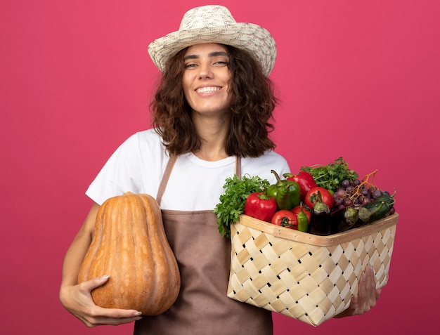 Sonriente joven jardinero en uniforme vistiendo sombrero de jardinería sosteniendo canasta de verduras con calabaza aislado en rosa