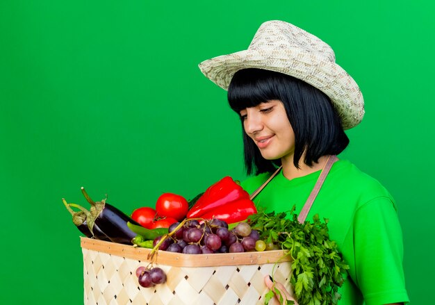 Sonriente joven jardinero en uniforme vistiendo sombrero de jardinería sosteniendo canasta de verduras