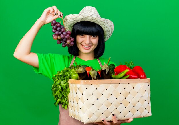 Sonriente joven jardinero en uniforme vistiendo sombrero de jardinería sosteniendo canasta de verduras
