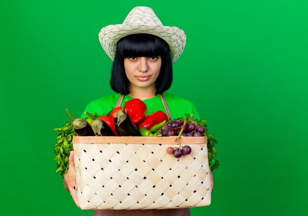 Sonriente joven jardinero en uniforme vistiendo sombrero de jardinería sosteniendo canasta de verduras