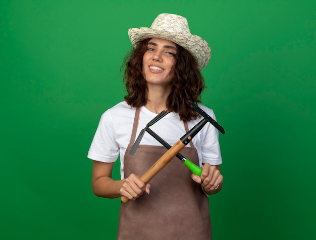 Sonriente joven jardinero en uniforme vistiendo sombrero de jardinería sosteniendo y cruzando rastrillo con rastrillo azada