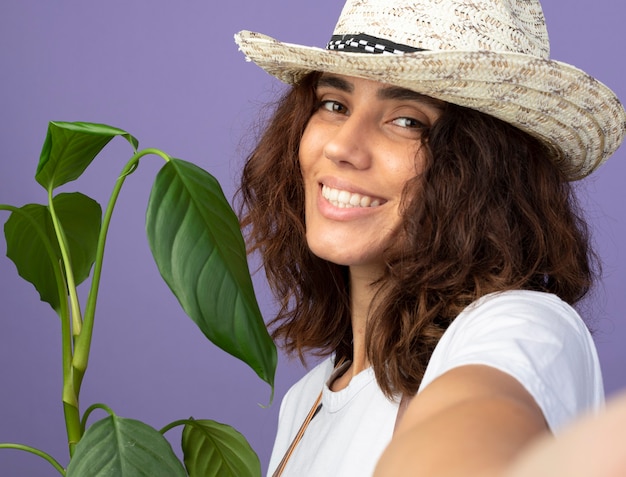 Sonriente joven jardinero en uniforme vistiendo sombrero de jardinería sosteniendo la planta