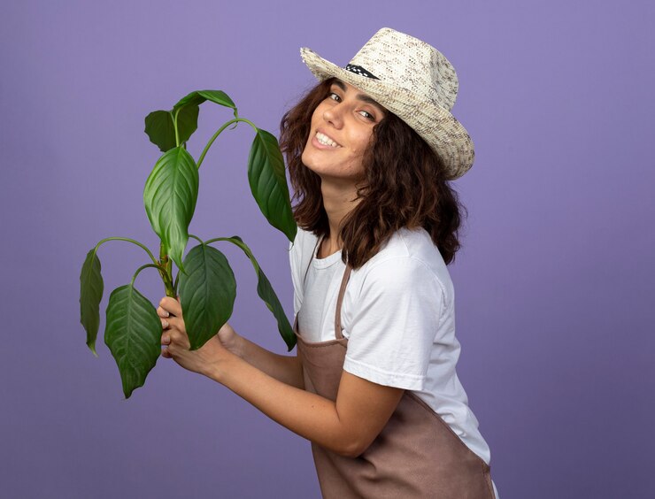 Foto gratuito sonriente joven jardinero en uniforme vistiendo sombrero de jardinería sosteniendo la planta