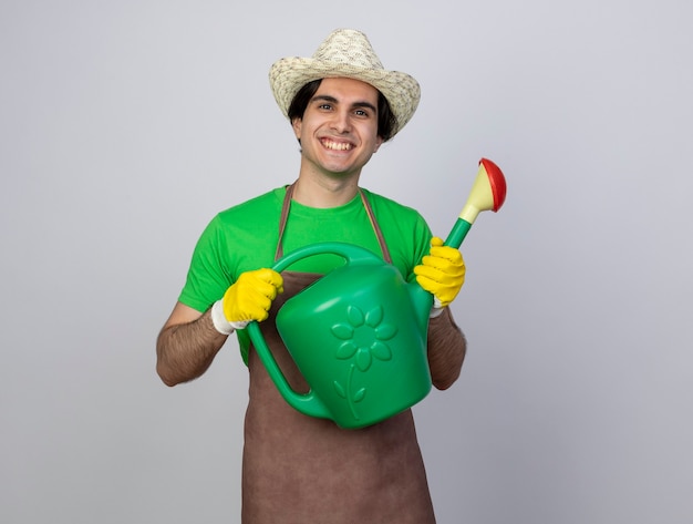 Sonriente joven jardinero en uniforme vistiendo sombrero de jardinería sosteniendo regadera