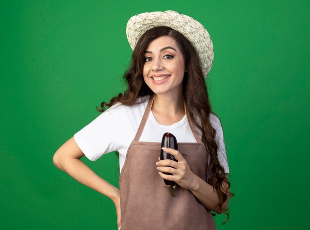 Sonriente joven jardinero en uniforme vistiendo sombrero de jardinería tiene berenjena aislado en la pared verde con espacio de copia