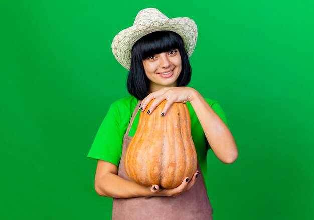 Sonriente joven jardinero en uniforme vistiendo sombrero de jardinería tiene calabaza mirando