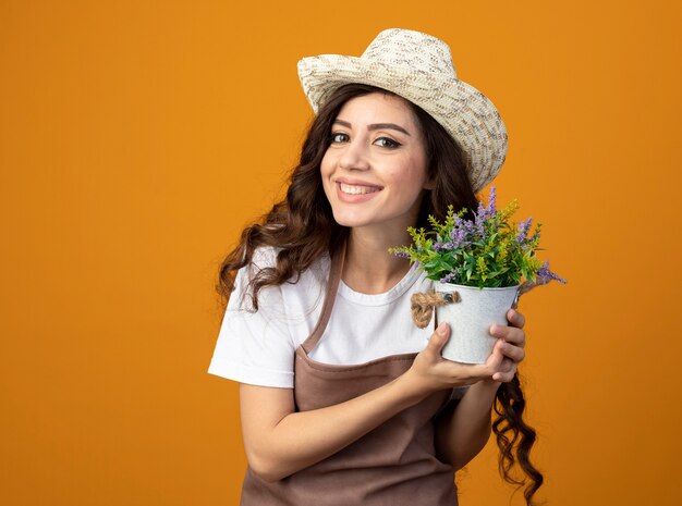 Sonriente joven jardinero en uniforme vistiendo sombrero de jardinería tiene maceta aislado en pared naranja con espacio de copia