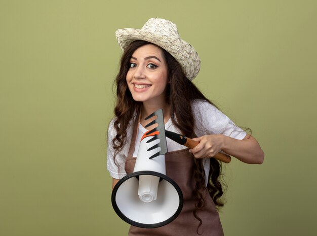 Sonriente joven jardinero en uniforme vistiendo sombrero de jardinería tiene rastrillo y altavoz aislado en la pared verde oliva