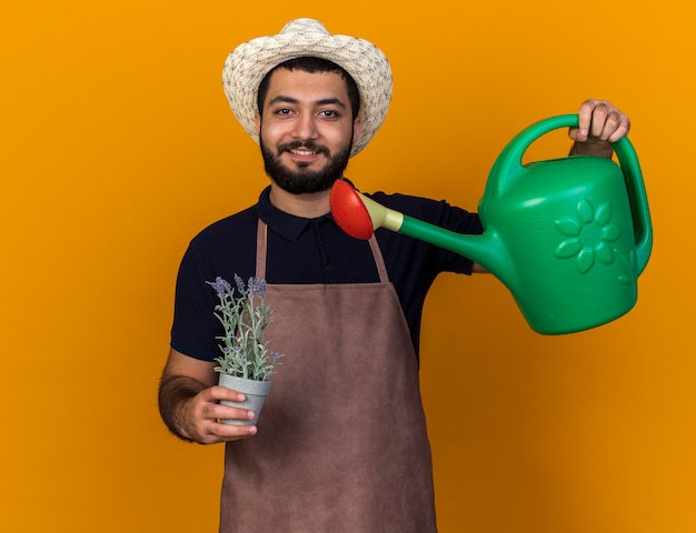 Foto gratuita sonriente joven jardinero varón caucásico con sombrero de jardinería fingiendo regar las flores en maceta con regadera aislado en la pared naranja con espacio de copia