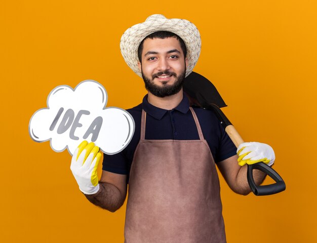 Sonriente joven jardinero varón caucásico vistiendo gorro y guantes de jardinería sosteniendo la burbuja de la idea y la pala en el hombro aislado en la pared naranja con espacio de copia