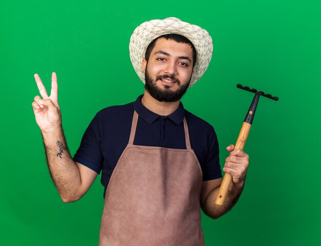 Sonriente joven jardinero varón caucásico vistiendo sombrero de jardinería sosteniendo el rastrillo y gesticulando el signo de la victoria aislado en la pared verde con espacio de copia