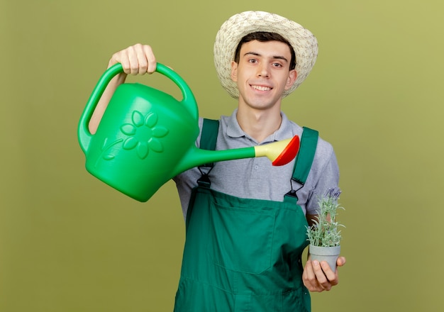 Sonriente joven jardinero vistiendo sombrero de jardinería pretende regar las flores