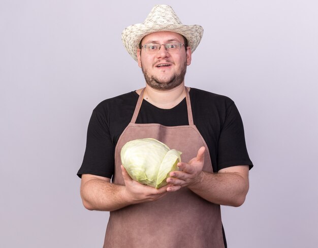 Sonriente joven jardinero vistiendo sombrero de jardinería sosteniendo repollo aislado en la pared blanca