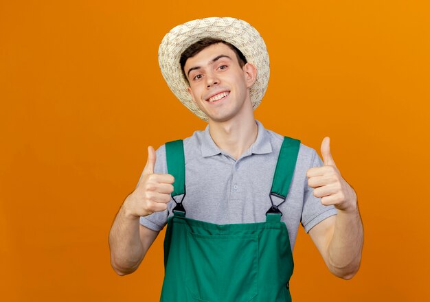 Sonriente joven jardinero vistiendo sombrero de jardinería Thumbs up con dos manos