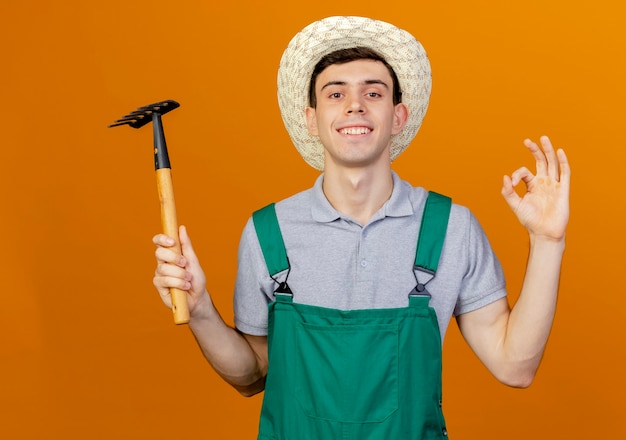 Sonriente joven jardinero vistiendo sombrero de jardinería tiene rastrillo y gestos ok signo de mano