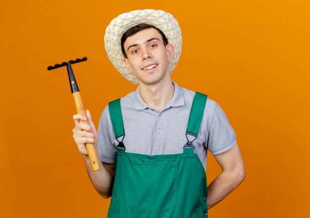 Sonriente joven jardinero vistiendo sombrero de jardinería tiene rastrillo